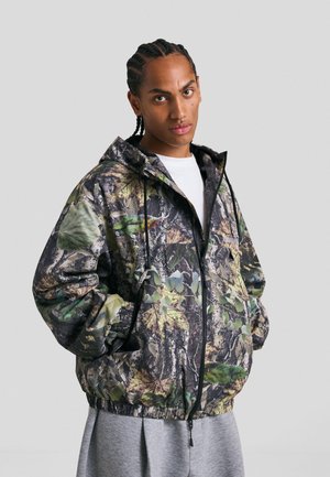 Giacca zip-up mimetica in tonalità di verde e marrone, con cappuccio e polsini elasticizzati, realizzata in materiale leggero.