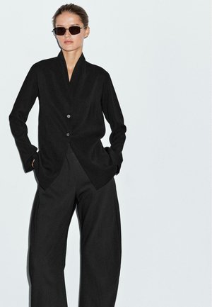Blazer nero con colletto a scialle, due bottoni e maniche lunghe. Abbinato a pantaloni neri larghi. Texture liscia, design minimalista.