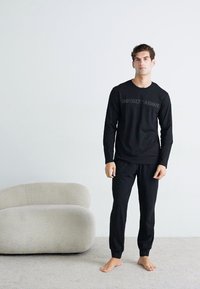 Čierny top s dlhým rukávom a detailom loga, spolu s nohavicami z mäkkej látky. Model stojí naboso vedľa zakriveného svetlého pohovky.