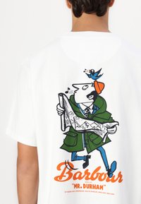 Valge T-särk, millel on graafikaprint rohelise mantli ja oranži mütsiga mehest, kes hoiab kätkeme suuri puid ja mägesid. Tekst: "Barbour 'Hr. Durham'."