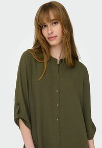 Camicia verde scuro con bottoni, colletto e maniche a tre quarti. Realizzata in tessuto morbido, presenta una finitura testurizzata e dettagli con bottoni.