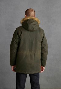 Olivově zelená parka s kožešinovou kapucí, s rovný střihem a dlouhými rukávy. Materiál vypadá texturovaně a je voděodolný.