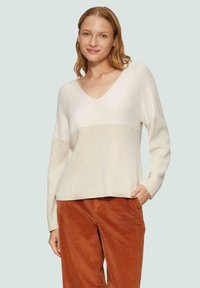 Pull en tricot crème avec un col en V, présentant une texture côtelée et une bande beige contrastante au milieu ; porté avec un pantalon en velours côtelé de couleur rouille.