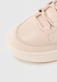 Primer plano de la puntera de una zapatilla beige con texturas lisas y de ante, costuras detalladas y cordones a juego sobre un fondo claro.