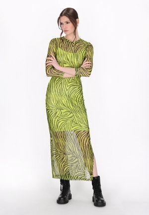 Femme debout, les bras croisés, portant une robe transparente à imprimé zèbre vert et noir et des bottes de combat noires sur fond blanc.