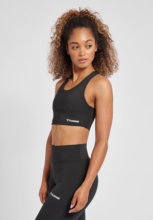 Mujer con pelo rizado que lleva un sujetador deportivo negro de Hummel y mallas de cintura alta, posando de perfil contra un fondo gris liso.