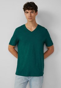 Koszulka z krótkim rękawem, w dekolcie w serek, w głębokim odcieniu teal, wykonana z lekkiego materiału o delikatnej teksturze. Dopasowany krój z metką na brzegu.