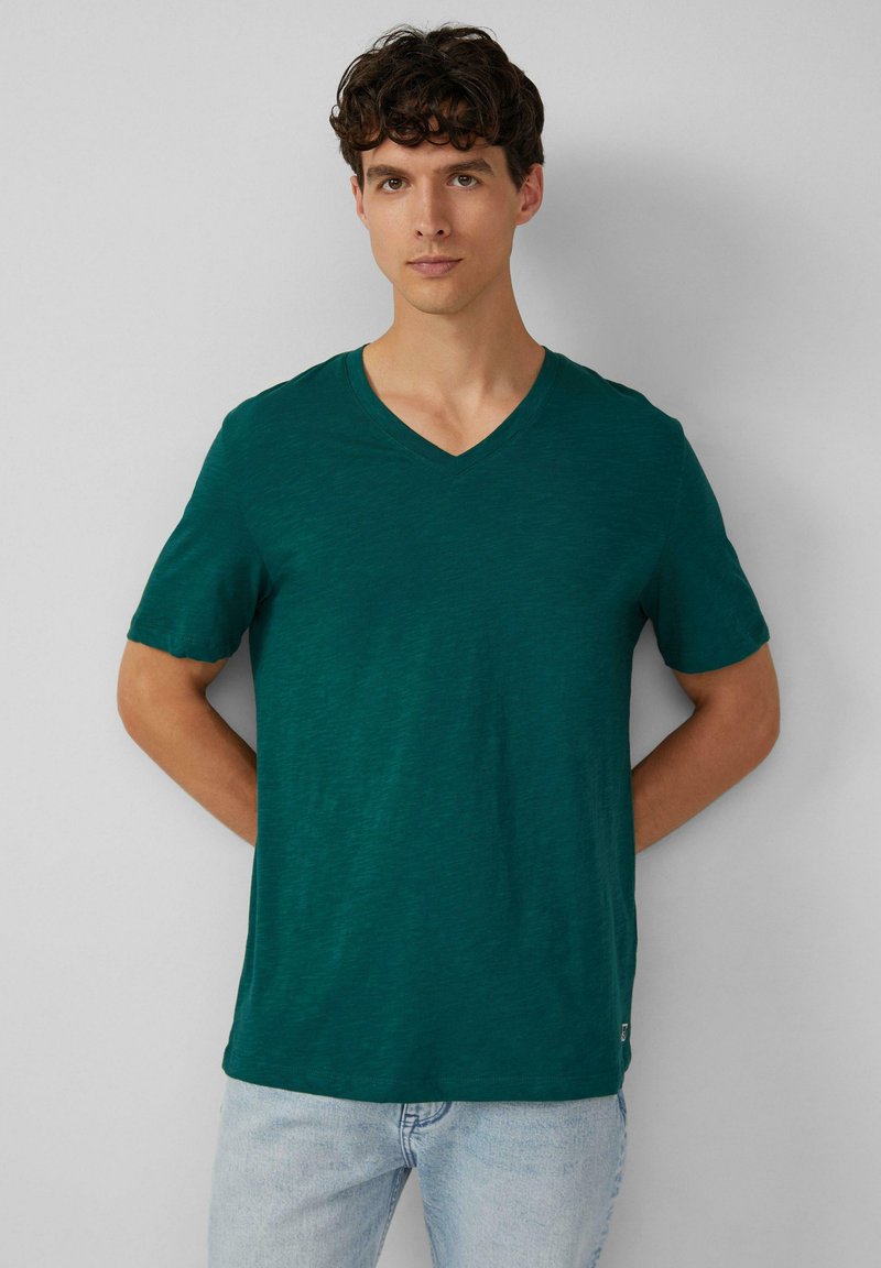 Koszulka z krótkim rękawem, w dekolcie w serek, w głębokim odcieniu teal, wykonana z lekkiego materiału o delikatnej teksturze. Dopasowany krój z metką na brzegu.