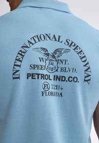 Ljusblå polo med svart tryckt text: "INTERNATIONAL SPEEDWAY," en örn-grafik och "PETROL IND. CO." med detaljer från Florida.