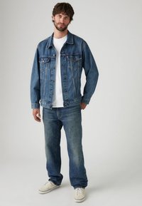 Jeans jakna srednje prane, z gumbi spredaj, dvema žepoma na prsih in sproščenega kroja. Oblečena z ohlapnimi kavbojkami in belimi supergami.