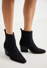 Svarta ankelstövlar i mjukt mocka, med spetsig tå, texturerade sidopaneler och en kraftig klack. Enkel design utan synlig hårdvara.