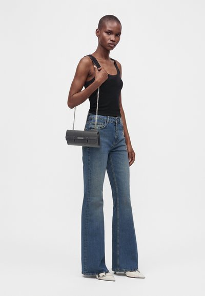 Femme aux cheveux courts portant un débardeur noir et un jean évasé bleu, tenant un petit sac noir avec une lanière chaîne sur l'épaule.