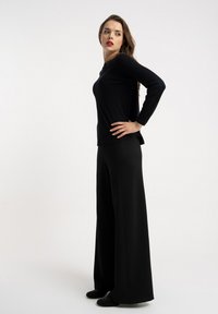 Czarna bluzka z długim rękawem w zestawieniu z szerokimi, czarnymi spodniami o luźnym kroju, oba wykonane z gładkiego materiału. Delikatne zdobienia na butach dodają subtelnej tekstury.