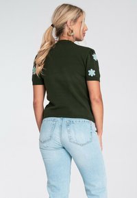 Kobieta z blond włosami związanymi w koński ogon, ubrana w ciemnozielony sweter z krótkim rękawem z niebieskimi wzorami kwiatowymi oraz jasnoniebieskie dżinsy, stojąca tyłem.