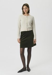 Cardigan beige clair avec des boutons, associé à une jupe vert foncé. Modèle portant des collants noirs et des ballerines à bout pointu.