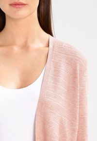Light pink cardigan made of ribbed material, featuring a relaxed fit and open front design, paired with a white tank top underneath.