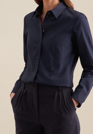 Kobieta w granatowej koszuli na guziki, wpuszczonej w spodnie w kolorze granatowym, plisowane, z wysokim stanem, ręce w kieszeniach.