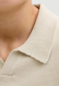 Col polo en tricot beige avec détail en tissu texturé, présentant un col arrondi et des coutures visibles, gros plan sur la zone du cou.
