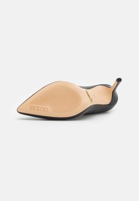 Svart spiss tå-sko med en glatt overflate og en subtil tekstur. Sålen er lys beige med riller og merket med "Guess."