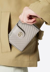 Gequilted grijze portemonnee met een textuur oppervlak, voorzien van een gouden logo accent en een drukknoopsluiting, gedeeltelijk zichtbaar in een beige jaszak.