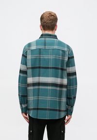 Flanelová košile v barvě tyrkysové, se vzorem černého a šedého kostkování. Dlouhé rukávy s knoflíkovými manžetami a mírně zakřivený lem. Měkká textura.