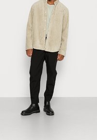 Camicia in corduroy beige con texture a costine, frontale bottonato e tasca. Abbinata a pantaloni neri a gamba stretta e stivaletti neri.
