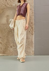 Dark brown cropped knit tank top with pink patterns, paired with loose beige trousers and light-colored slip-on sandals. Beret-style bag held.