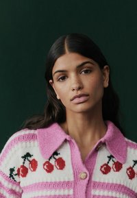 Gebreid vest met roze kraag en knoop, met rode kersen op een witte achtergrond, met horizontale roze strepen en zachte textuur.