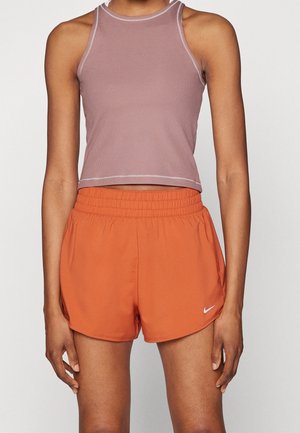 Kvinde iført ærmeløs lilla crop top og orange Nike sportsshorts med elastisk talje, stående mod en ensfarvet baggrund.