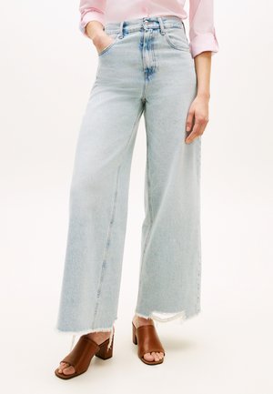 Frau trägt hellblaue Jeans mit weitem Bein und ausgefransten Säumen, ein rosafarbenes hochgekrempeltes Hemd und braune offene Sandalen mit Absatz, steht mit den Händen in den Taschen.