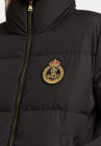 Mustat toppatakki, jossa on kultainen vetskari ja kirjaillut vaakuna-ompelu, jossa on kruunu ja laakerinlehdet vasemmassa rinnassa.