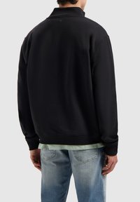 Zwarte pulloverjack met hoge kraag, geribbelde manchetten en een relaxte pasvorm, gedragen over een lichtgro shirt, gecombineerd met lichtblauwe jeans.