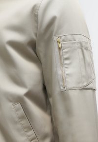 Beige jas met een mouw met een zak met ritssluiting en extra stikdetails op de bovenarm.