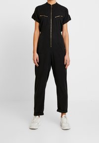 Zwarte jumpsuit met korte omgerolde mouwen, gouden voorrits, borstzakken en loszittende benen. Draag het met witte sneakers.
