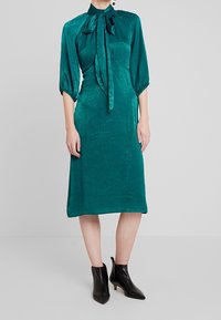 Teal sident klänning med hög krage, fluga och trekvarts puffärmar. Figurnära livstycke och flödande midiklänning, kombinerad med svarta ankelboots.