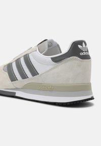 Vita och grå Adidas-sneakers med mockadetaljer, tre grå ränder och Adidas-logotyp på hälen och mellansulan, sedd från baksidan.
