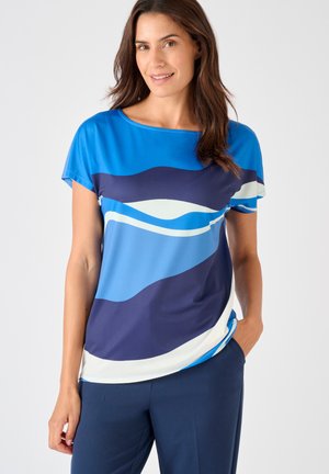 Donna che indossa una maglietta blu a maniche corte con motivi astratti a onde in blu navy, azzurro e bianco, abbinata a pantaloni blu scuro, in piedi con una mano in tasca.