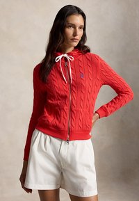 Vrouw die een rood kabelgebreid hoodie met een ritssluiting draagt, met witte trekkoorden en witte shorts, poserend tegen een neutrale achtergrond.