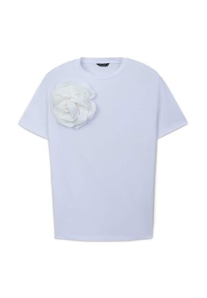 T-shirt en coton blanc à manches courtes, arborant un grand accent en forme de fleur en tissu blanc sur le côté gauche. Texture lisse et coupe ample.