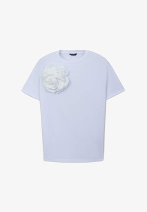 T-shirt bianco in cotone con maniche corte, caratterizzato da un grande fiore in tessuto bianco applicato sul lato sinistro. Texture liscia e vestibilità ampia.