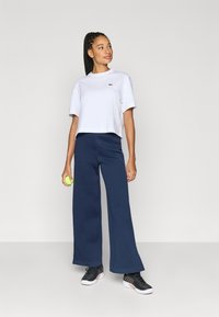 Λευκό κοντό μπλουζάκι με μικρό πράσινο λογότυπο, συνδυασμένο με μπλε σκούρο παντελόνι φαρδιάς γραμμής. Το μοντέλο κρατά μια πράσινη μπάλα του τένις και φορά μαύρα αθλητικά παπούτσια.