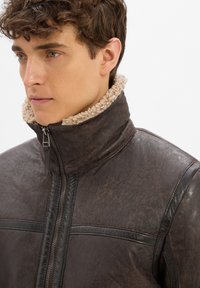 Brązowa skórzana kurtka o teksturowanym wykończeniu, z wysokim kołnierzem wyłożonym beżowym sztucznym futrem i metalowym zapięciem na zamek.