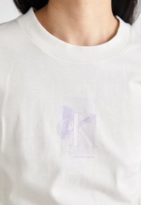 Biała bawełniana koszulka z okrągłym dekoltem, z fioletowym logo graficznym "CK" i napisem "Calvin Klein Jeans" poniżej. Miękka w dotyku.