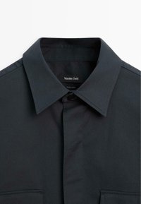 Dark gray button-up shirt with a classic collar, made from smooth fabric. Features a front pocket and minimal stitching details.