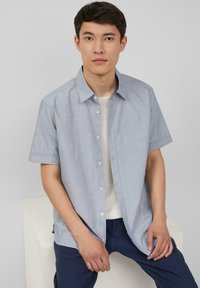 Kurzarm-Hemd mit Knopfleiste aus hellblauem Stoff mit dezentem Muster, kombiniert mit einem weißen Rundhals-T-Shirt und dunkelblauen Hosen.