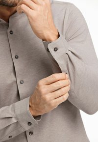 Camicia grigia chiara con colletto alla coreana, con bottoni neri visibili e un polsino arrotolato. Texture del tessuto liscia.