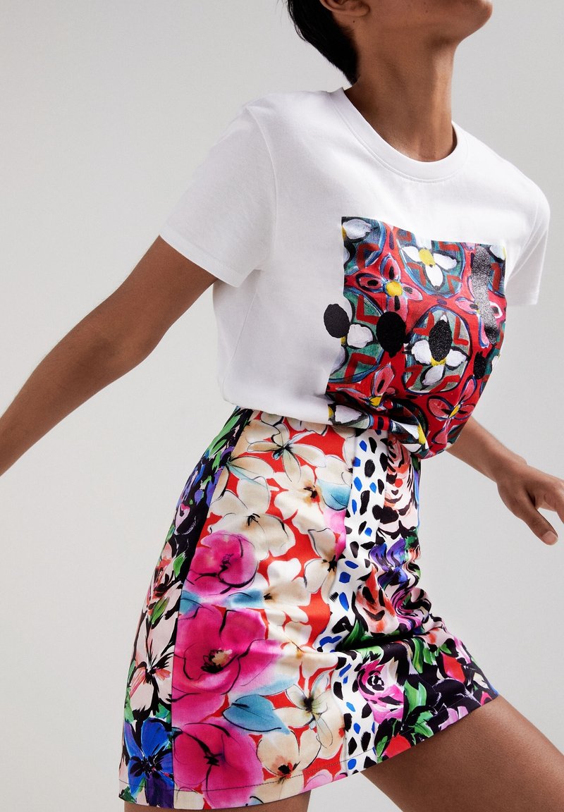Personne portant un t-shirt blanc avec un imprimé floral abstrait et coloré, ainsi qu'une jupe à motif floral vibrant dans des tons rouges, roses, bleus et verts.