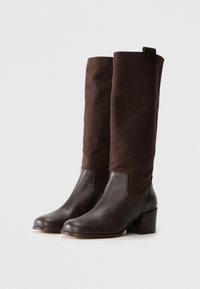 Une paire de bottes marron foncé montant jusqu'au genou avec une tige en cuir et des talons blocs, un empattement en tissu et une languette de tirage en haut.
