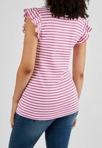 Różowo-biała bluzka w paski z falbanami na rękawach, dopasowany krój i zaokrąglony dół, w zestawieniu z niebieskimi dżinsami.