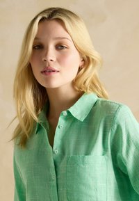 Groene, textuurvolle button-up overhemd met een kraag, een enkele borstzak en een lichte knoopontwerp, tegen een neutrale achtergrond.